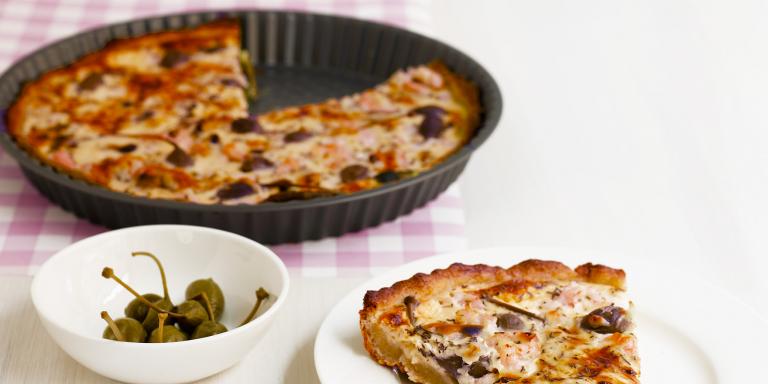 Pyöreä piirakkavuoka, josta otettu piirakkapala lautaselle, kapriksia kipossa.