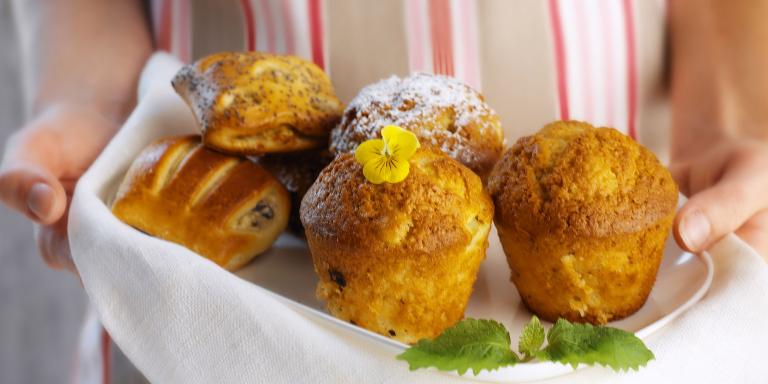 Kädet pitelevät tarjotinta jossa on vaaleita muffinsseja ja muita leivonnaisia