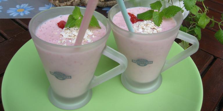 Vaaleanpunainen mustikkasmoothie tarjoiltuna kahdesta huurrelasisesta kahvikupista pilleineen, koristeltu punaisilla marjoilla, kaurahiutaleilla ja sitruunamelissan lehdillä.