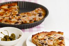 Pyöreä piirakkavuoka, josta otettu piirakkapala lautaselle, kapriksia kipossa.