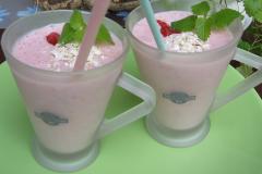 Vaaleanpunainen mustikkasmoothie tarjoiltuna kahdesta huurrelasisesta kahvikupista pilleineen, koristeltu punaisilla marjoilla, kaurahiutaleilla ja sitruunamelissan lehdillä.